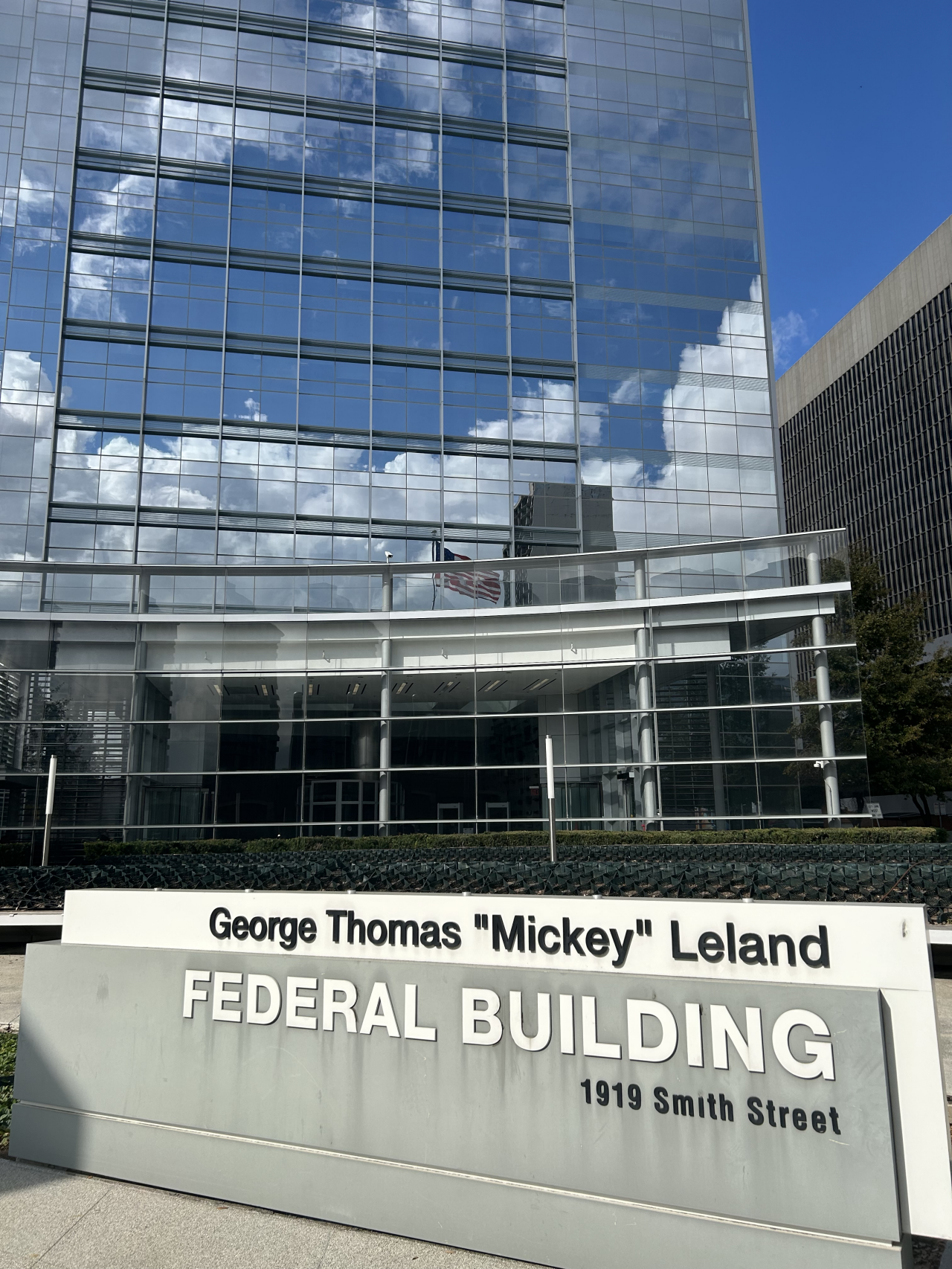 Exterior of the Houston Passport Agency. It is located in a tall office building with mirrored windows. It sits behind a sign that says “George Thomas ‘Mickey’ Leland FEDERAL BUILDING 1919 Smith Street”.